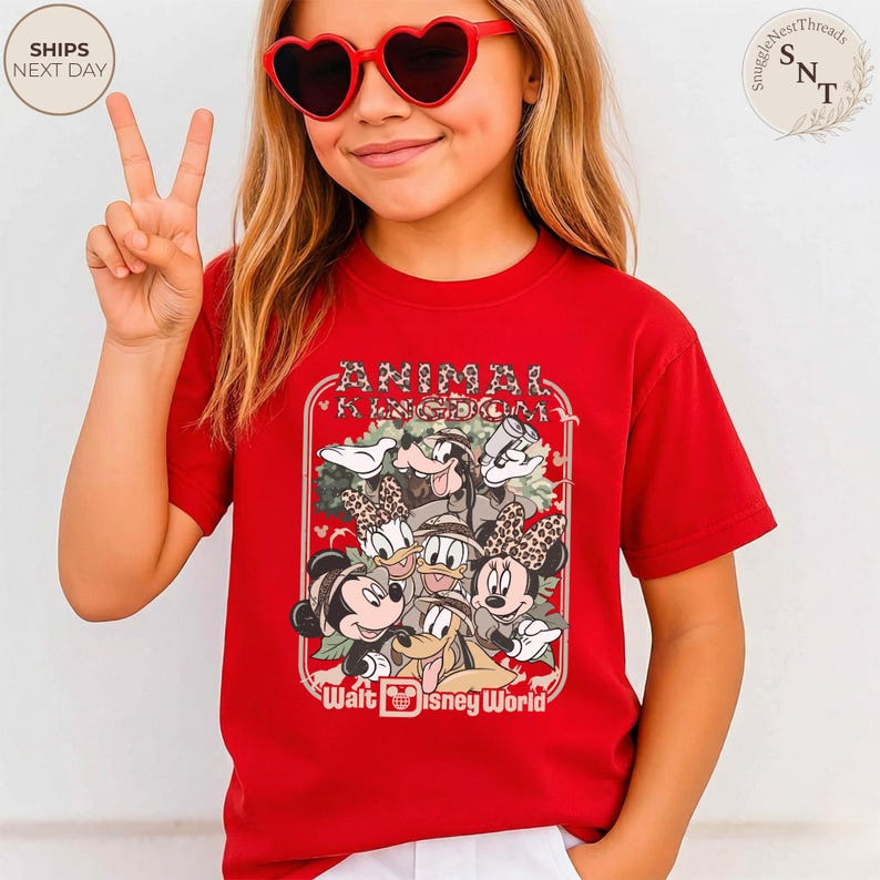 Pu&ograve; includere: Una maglietta rossa con una grafica di personaggi Disney, tra cui Topolino e Minnie, Paperino e Paperina, e Pippo. La maglietta presenta la scritta "Animal Kingdom" e "Walt Disney World" in una cornice decorativa.