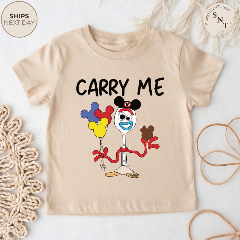 May include: A beige short-sleeved t-shirt featuring the phrase "CARRY ME" in black letters. The shirt has a cartoon graphic of a fork with Mickey Mouse ears, holding balloons and a chocolate treat. The shirt is designed for children.