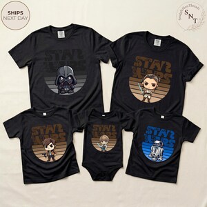 May include: Black t-shirts with Star Wars graphics featuring Darth Vader, Rey, Han Solo, a baby with a lightsaber, and R2-D2. The shirts have a retro design with a brown and blue color scheme.