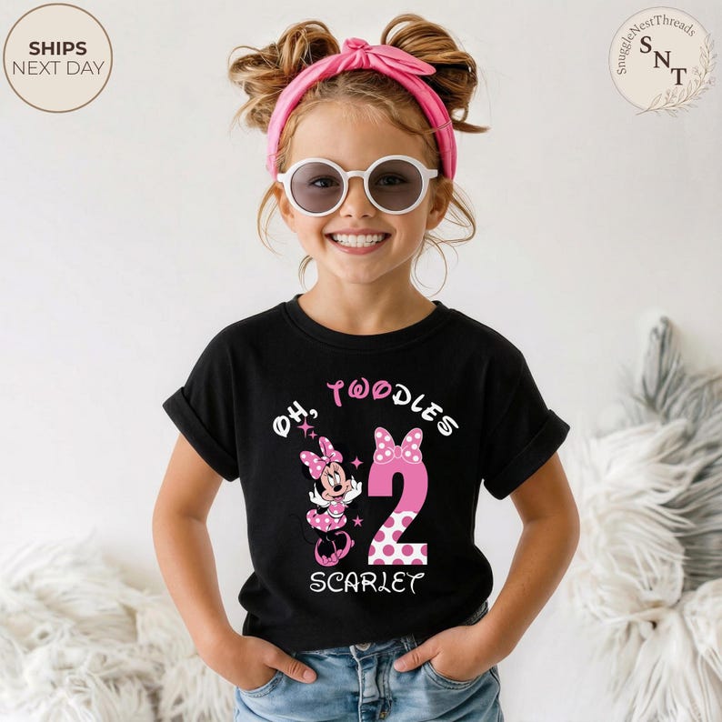 Puede incluir: Una camiseta negra con un dise&ntilde;o de Minnie Mouse rosa y el texto "Oh, Twoodles 2 Scarlet". El n&uacute;mero 2 es rosa con lunares blancos. La ni&ntilde;a lleva gafas de sol y una diadema rosa.