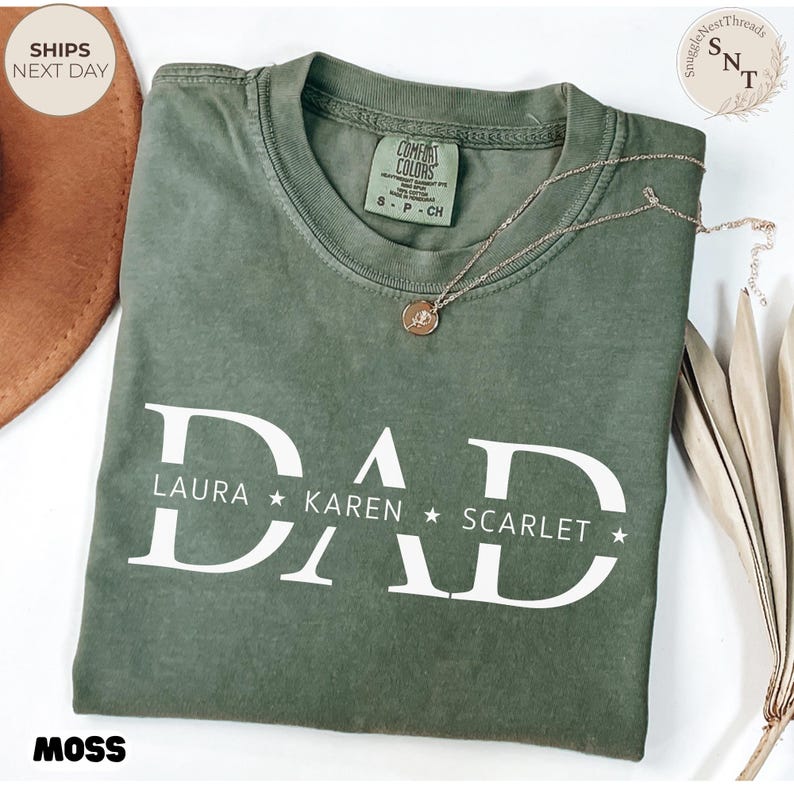 Puede incluir: Camiseta verde musgo con la palabra "DAD" en letras blancas grandes, con los nombres LAURA, KAREN y SCARLET debajo. Un collar dorado con un colgante descansa sobre la camiseta. La camiseta est&aacute; etiquetada como "Comfort Colors" y talla S-P-CH.