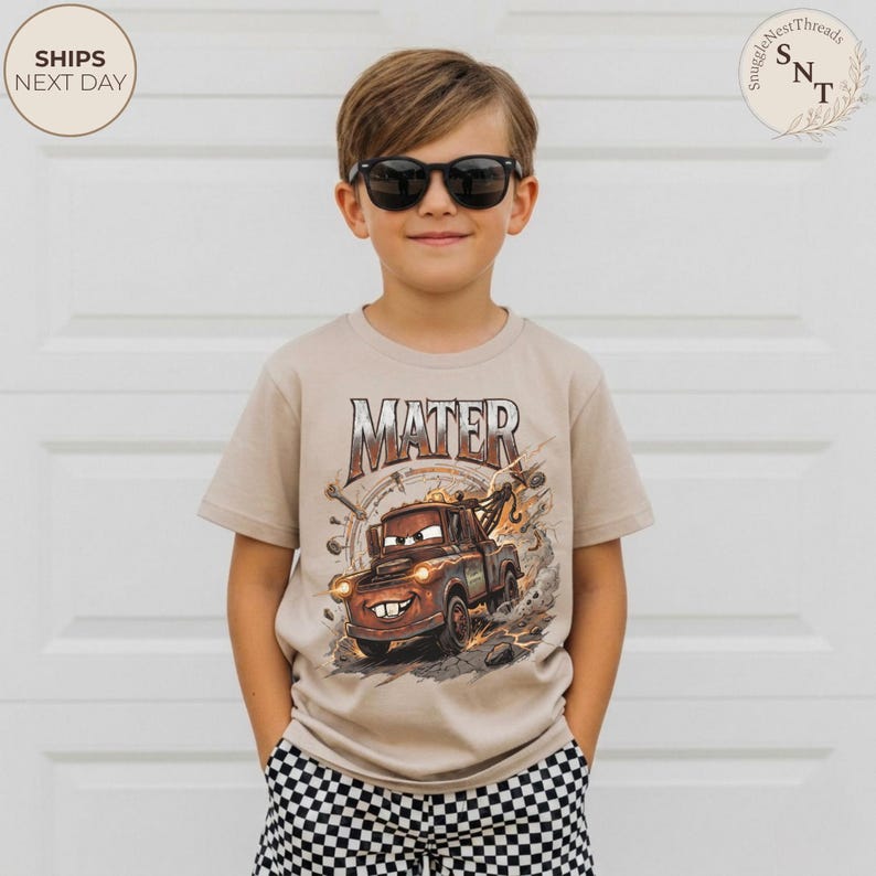 Puede incluir: Una camiseta beige de manga corta con un gr&aacute;fico del personaje Mater de la pel&iacute;cula Cars. La palabra "MATER" est&aacute; impresa encima de la imagen. El ni&ntilde;o lleva gafas de sol negras y pantalones cortos a cuadros.