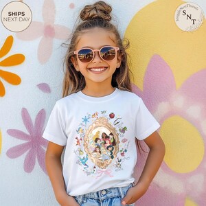 May include: A white t-shirt with a gold-framed illustration of Disney princesses. The design includes various characters and elements from Disney movies. The girl is wearing pink sunglasses and is smiling. The background is a colourful mural.