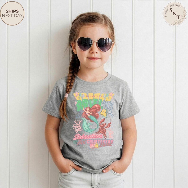 May include: A heather gray short-sleeve t-shirt featuring a colorful graphic print of a mermaid, a crab, and a fish. The text on the shirt reads "ATLANTIC ARIEL OCEAN TOUR 1991" and "Sebastian and Crustacean Band". The shirt has a crew neckline.