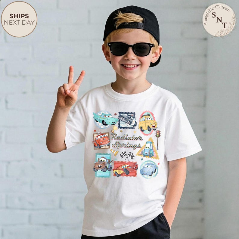 May include: White t-shirt featuring colorful cartoon car designs and the text "Radiator Springs". The shirt is worn by a child wearing sunglasses and a black cap.