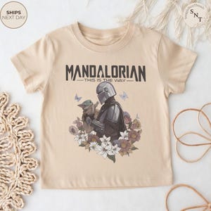 Puede incluir: Una camiseta beige de manga corta con un gr&aacute;fico del Mandaloriano y Grogu rodeados de flores y mariposas. El texto "MANDALORIAN" y "THIS IS THE WAY" est&aacute; impreso en negro sobre los personajes con una tipograf&iacute;a negrita y estilizada.
