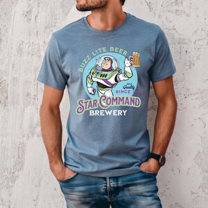 Puede incluir: Camiseta azul claro con una ilustraci&oacute;n de dibujos animados de Buzz Lightyear sosteniendo una jarra de cerveza. El dise&ntilde;o incluye el texto "Buzz Lite Beer" y "Star Command Brewery" en un estilo vintage. La camiseta es de material suave.