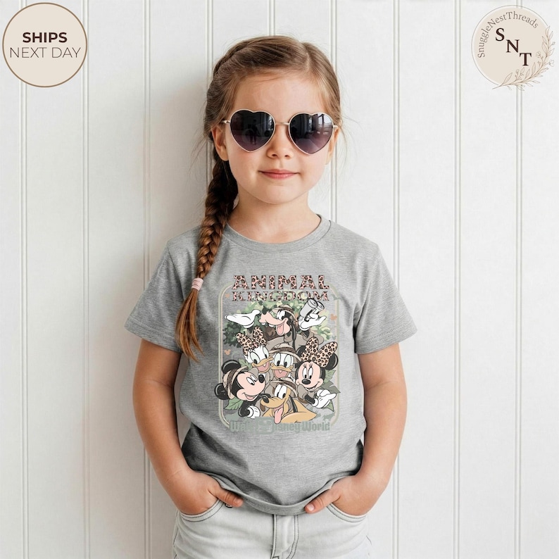 Pu&ograve; includere: Una maglietta grigia con una grafica di Topolino, Minnie, Paperino e Pluto, con la scritta "Animal Kingdom" e "Where the Wild Things Roam". La bambina indossa occhiali da sole a forma di cuore.