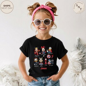 May include: A black t-shirt featuring a colorful graphic of Disney characters dressed as Avengers. The shirt includes Mickey Mouse, Minnie Mouse, Donald Duck, and others. The word "Avengers" is printed in red below the characters.