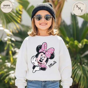 May include: A white sweatshirt featuring a Minnie Mouse graphic with a pink polka-dot bow. The word "Minnie" is written in cursive. The sweatshirt is worn by a young person wearing a black beanie and sunglasses.
