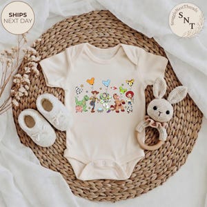 May include: Cream-colored baby onesie featuring a colorful graphic of Toy Story characters. The design includes Woody, Buzz Lightyear, and other characters. The onesie is placed on a woven wicker mat with a pair of baby shoes and a stuffed bunny.