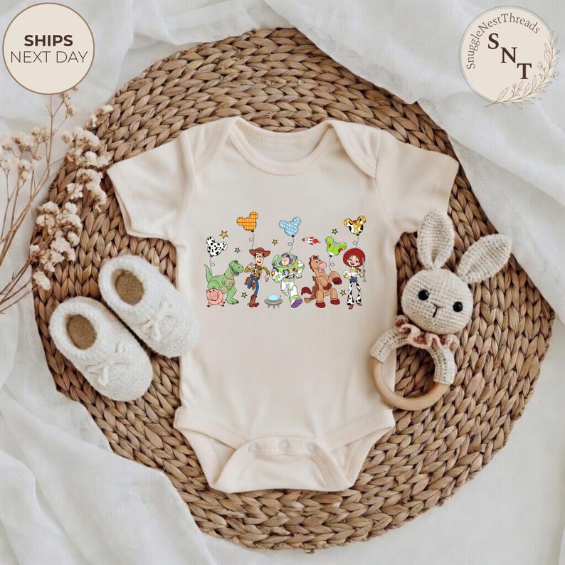 May include: Cream-colored baby onesie featuring a colorful graphic of Toy Story characters. The design includes Woody, Buzz Lightyear, and other characters. The onesie is placed on a woven wicker mat with a pair of baby shoes and a stuffed bunny.