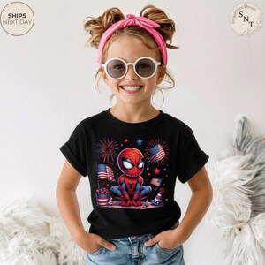 May include: A black short-sleeve t-shirt featuring a graphic of a cartoon spider-themed character in a crouched pose, surrounded by red, white, and blue fireworks, American flags, and festive party hats, worn with blue denim jeans.