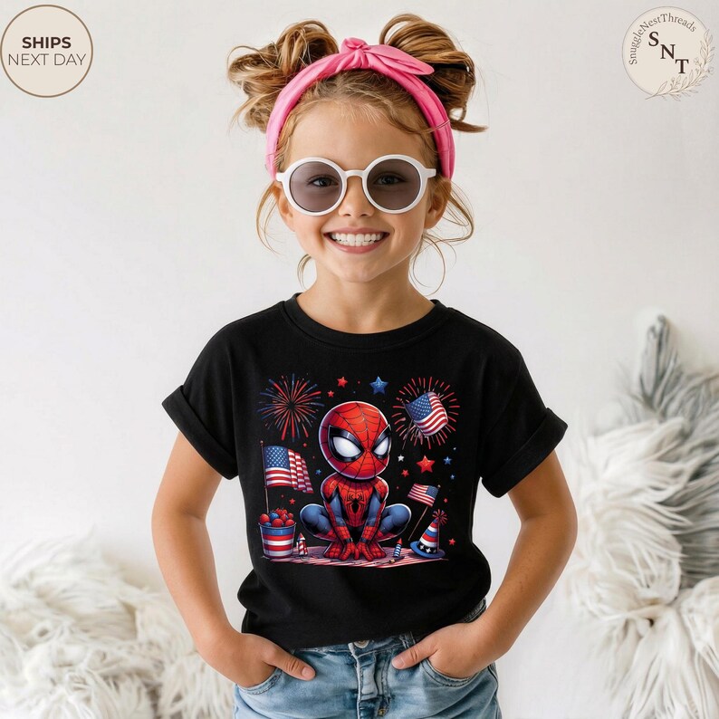 May include: A black short-sleeve t-shirt featuring a graphic of a cartoon spider-themed character in a crouched pose, surrounded by red, white, and blue fireworks, American flags, and festive party hats, worn with blue denim jeans.
