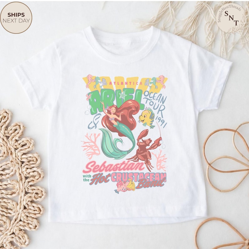 May include: A white short-sleeved t-shirt featuring a colorful graphic print of Ariel, Flounder, and Sebastian. The text reads Atlantica Ocean Tour 1991, Sebastian with the Hot Crustacean Band, and Ariel. The design includes coral and bubble motifs.