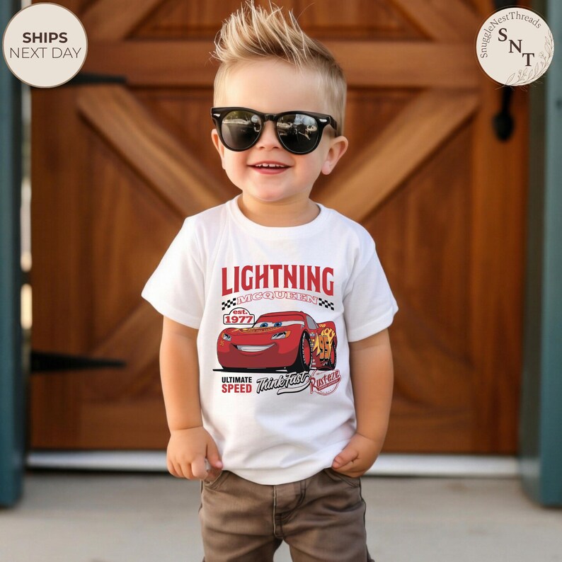 Op de afbeelding: Een wit T-shirt met een afbeelding van Bliksem McQueen uit de film Cars. Het ontwerp bevat de tekst "LIGHTNING MCQUEEN" in het rood, het jaar "1977" en de zinnen "ULTIMATE SPEED" en "Think Fast". Het T-shirt wordt door een jong kind gedragen.