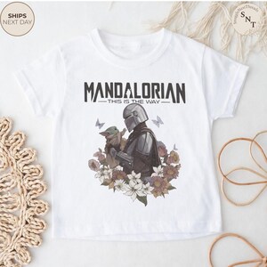 Puede incluir: Una camiseta blanca de manga corta con un estampado gr&aacute;fico del Mandaloriano sosteniendo a Grogu, rodeado por un grupo de flores coloridas y mariposas. El texto "MANDALORIAN THIS IS THE WAY" est&aacute; impreso en negro sobre la ilustraci&oacute;n.