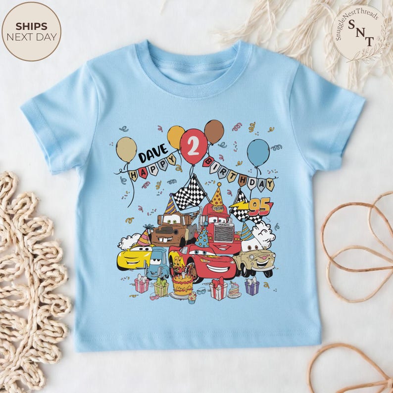 Puede incluir: Camiseta infantil azul claro con un dise&ntilde;o colorido de coches de dibujos animados celebrando un cumplea&ntilde;os. El dise&ntilde;o incluye globos, confeti, una tarta de cumplea&ntilde;os y el texto "DAVE HAPPY BIRTHDAY 2".