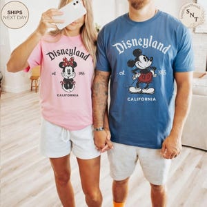 Pu&ograve; includere: Coppia di magliette Disneyland rosa e blu con grafiche di Topolino e Minnie. Le magliette presentano la scritta "Disneyland", "est. 1955" e "California". La coppia indossa pantaloncini grigi. L'immagine &egrave; stata scattata in casa.