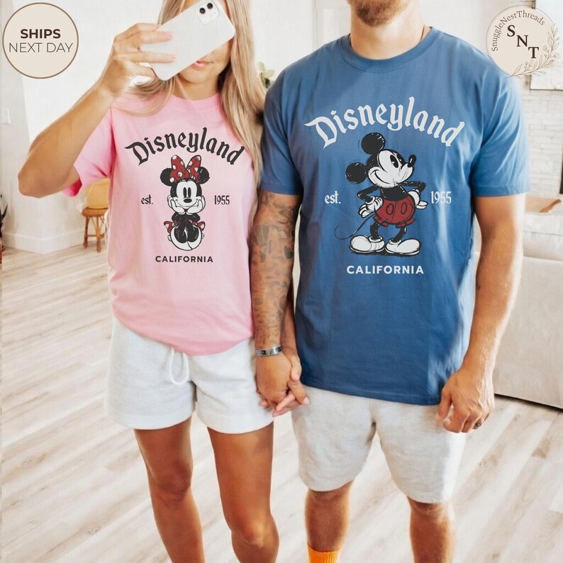Pu&ograve; includere: Coppia di magliette Disneyland rosa e blu con grafiche di Topolino e Minnie. Le magliette presentano la scritta "Disneyland", "est. 1955" e "California". La coppia indossa pantaloncini grigi. L'immagine &egrave; stata scattata in casa.
