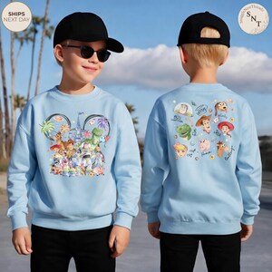 Puede incluir: Una sudadera azul claro con un gr&aacute;fico frontal de personajes de Toy Story dentro de una silueta de orejas de Mickey Mouse y un gr&aacute;fico trasero con las caras de Buzz Lightyear, Woody, Jessie, Rex y Hamm con sus nombres escritos en cursiva.