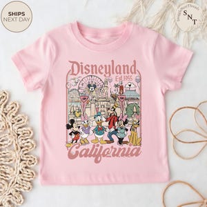 May include: A light pink short-sleeve t-shirt featuring a graphic print of Disneyland California with the text Disneyland Est. 1955 California. The illustration includes Mickey Mouse, Minnie Mouse, Donald Duck, Daisy Duck, Goofy, and Pluto.
