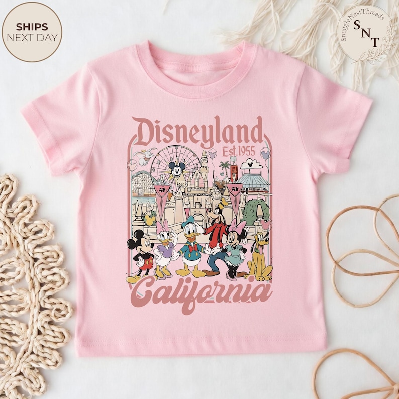 May include: A light pink short-sleeve t-shirt featuring a graphic print of Disneyland California with the text Disneyland Est. 1955 California. The illustration includes Mickey Mouse, Minnie Mouse, Donald Duck, Daisy Duck, Goofy, and Pluto.