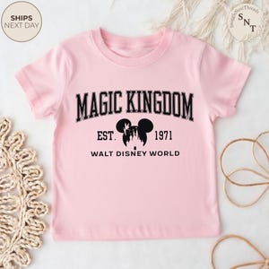 May include: A light pink t-shirt featuring the words "MAGIC KINGDOM" in black block letters. Below the text is a silhouette of Mickey Mouse ears and a castle, with "EST. 1971" and "WALT DISNEY WORLD" underneath.