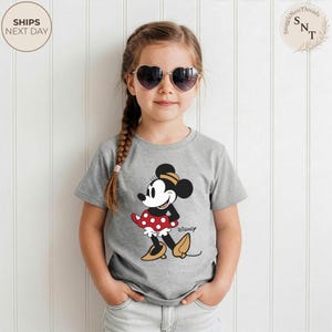 May include: A gray t-shirt featuring a Minnie Mouse graphic. The cartoon character is depicted in a red polka-dot skirt, black top, and yellow shoes. The word "Disney" is printed below the graphic.