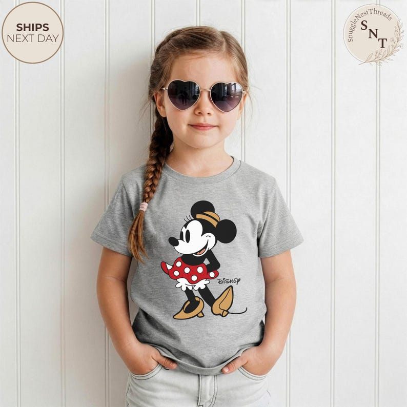 May include: A gray t-shirt featuring a Minnie Mouse graphic. The cartoon character is depicted in a red polka-dot skirt, black top, and yellow shoes. The word "Disney" is printed below the graphic.