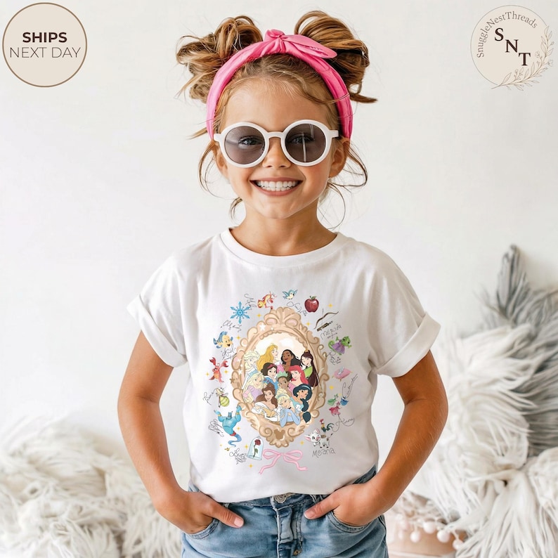 May include: A white t-shirt with a gold-framed portrait of Disney princesses and cartoon illustrations. The girl is wearing sunglasses and a pink headband. The shirt design includes characters like Cinderella, Snow White, and Belle.