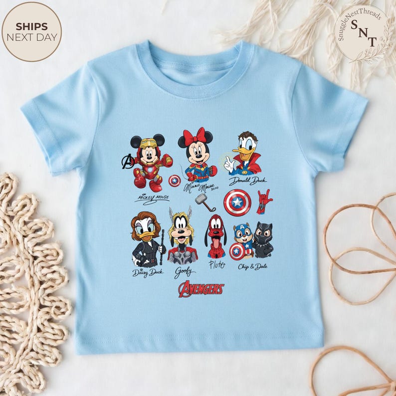 May include: A light blue t-shirt featuring a colorful graphic of various Disney and Marvel characters, including Mickey Mouse, Minnie Mouse, and Donald Duck. The word "AVENGERS" is printed in red at the bottom. The shirt is made of soft material.
