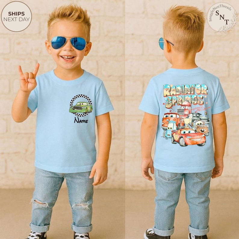 May include: Light blue t-shirt featuring a green race car graphic inside a checkered circle, with the word "Name" below. The back of the shirt displays "Radiator Springs" text and cartoon car characters. The child is wearing blue sunglasses and jeans.