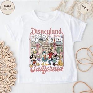 May include: A white short-sleeve t-shirt featuring a graphic print of Disneyland California with the text Disneyland Est. 1955 California. The illustration includes Mickey Mouse, Minnie Mouse, Donald Duck, Daisy Duck, Goofy, and Pluto.