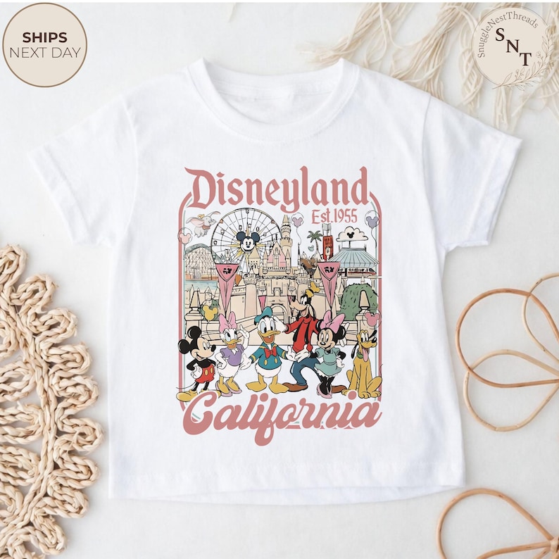 May include: A white short-sleeve t-shirt featuring a graphic print of Disneyland California with the text Disneyland Est. 1955 California. The illustration includes Mickey Mouse, Minnie Mouse, Donald Duck, Daisy Duck, Goofy, and Pluto.