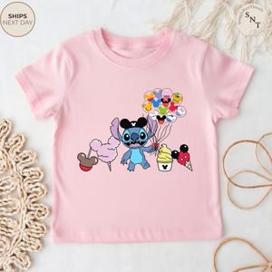 Pu&ograve; includere: T-shirt rosa chiaro con un personaggio dei cartoni animati di Stitch con orecchie di Topolino e palloncini. Il design include zucchero filato, gelato e un cupcake. La maglietta ha girocollo e maniche corte.