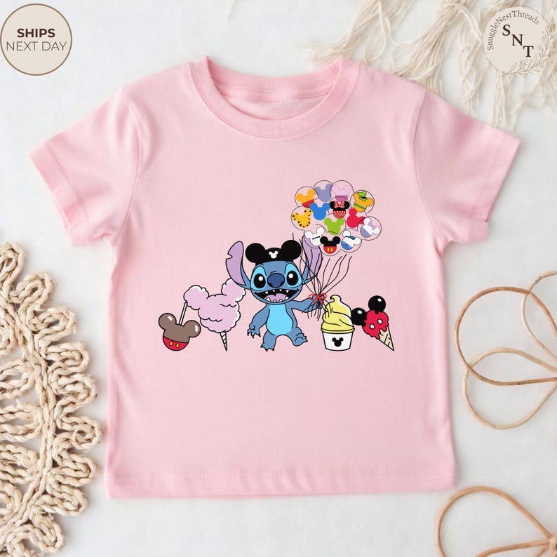 Pu&ograve; includere: T-shirt rosa chiaro con un personaggio dei cartoni animati di Stitch con orecchie di Topolino e palloncini. Il design include zucchero filato, gelato e un cupcake. La maglietta ha girocollo e maniche corte.