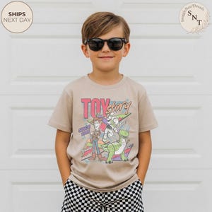 Może przedstawiać: Beżowa koszulka z kr&oacute;tkim rękawem z kolorową grafiką "Toy Story" z Woodym, Buzzem Astralem i Rexem. Tekst zawiera "TOY STORY" i "SINCE 1995". Chłopiec ma na sobie czarne okulary przeciwsłoneczne i szorty w kratę.