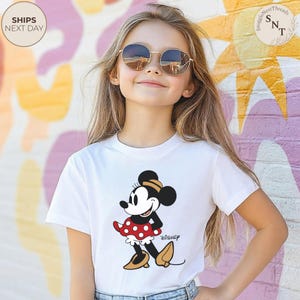 May include: A white t-shirt featuring a cartoon Minnie Mouse design. Minnie Mouse is depicted in a red polka dot dress, black top, and yellow shoes. The word "Disney" is printed below the character.