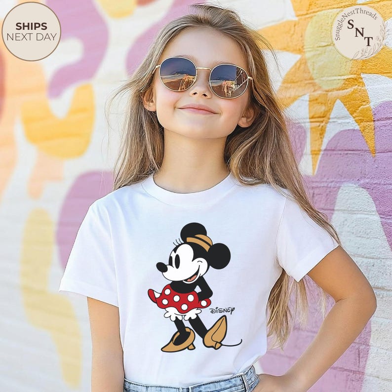 May include: A white t-shirt featuring a cartoon Minnie Mouse design. Minnie Mouse is depicted in a red polka dot dress, black top, and yellow shoes. The word "Disney" is printed below the character.