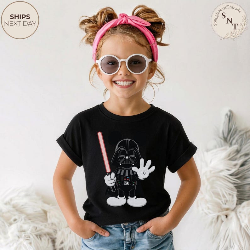 May include: Black t-shirt featuring a cartoon Darth Vader character holding a red lightsaber and waving. The graphic is in a cartoon style. The shirt has rolled-up sleeves. The text "SHIPS NEXT DAY" is in a circle.