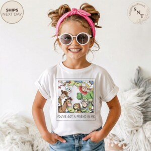 May include: A white t-shirt featuring a colorful graphic of characters from the movie "Toy Story." The shirt has the text "YOU'VE GOT A FRIEND IN ME" at the bottom. The child is wearing sunglasses and a pink headband.