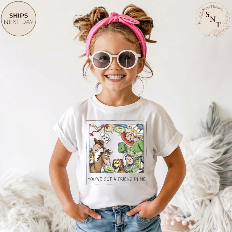 May include: A white t-shirt featuring a colorful graphic of characters from the movie "Toy Story." The shirt has the text "YOU'VE GOT A FRIEND IN ME" at the bottom. The child is wearing sunglasses and a pink headband.