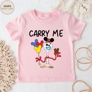 May include: A light pink t-shirt featuring a cartoon character holding balloons and a chocolate treat. The text "CARRY ME" is printed in black. The character has a spoon for a body and a fork for legs.