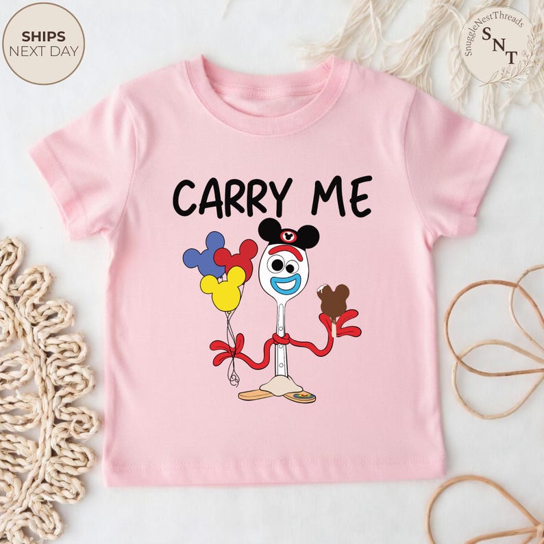 May include: A light pink t-shirt featuring a cartoon character holding balloons and a chocolate treat. The text "CARRY ME" is printed in black. The character has a spoon for a body and a fork for legs.