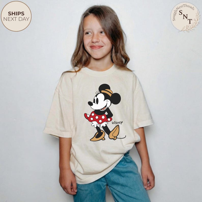 May include: A cream-colored t-shirt featuring a graphic of Minnie Mouse in a red polka-dot dress, black shoes, and a yellow hat. The Disney logo is printed below the character. The shirt is worn by a person wearing blue jeans.