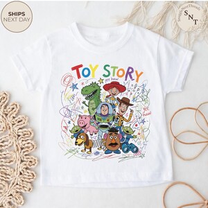 May include: White t-shirt featuring a colorful graphic of characters from the animated film "Toy Story." The design includes the words "Toy Story" in vibrant colors, along with images of Woody, Buzz Lightyear, and other characters.