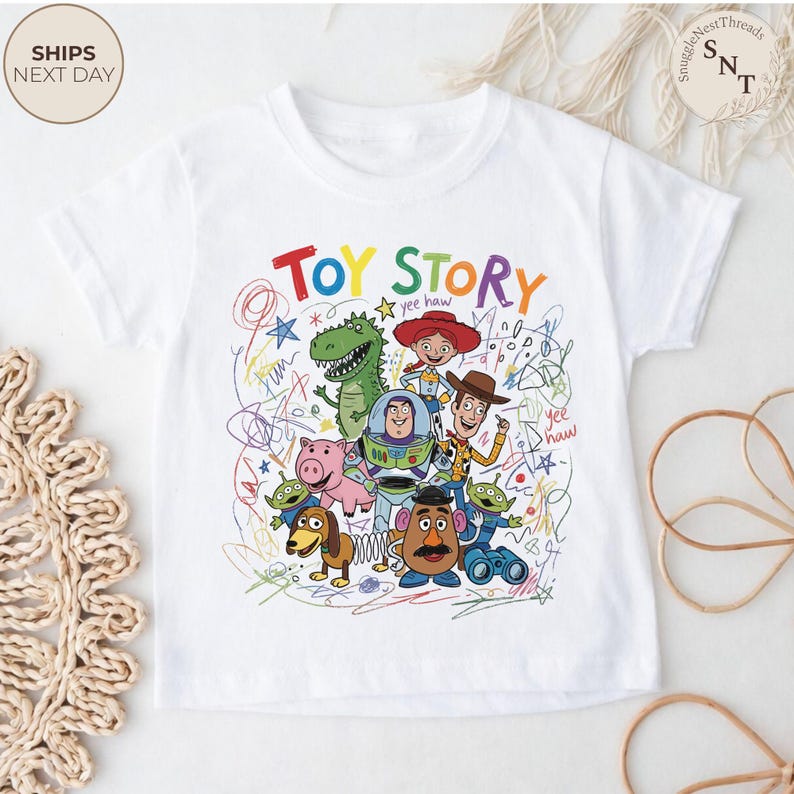 May include: White t-shirt featuring a colorful graphic of characters from the animated film "Toy Story." The design includes the words "Toy Story" in vibrant colors, along with images of Woody, Buzz Lightyear, and other characters.