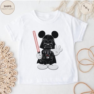 May include: White short-sleeved t-shirt featuring a cartoon illustration of Darth Vader with Mickey Mouse ears. The character is holding a red lightsaber and waving. The shirt has the text "SnuggleNestThreads" and "Ships Next Day".