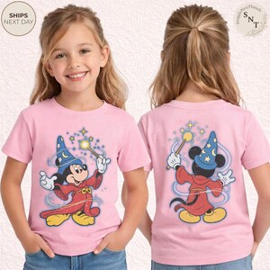 Op de afbeelding: Roze T-shirts met een cartoonafbeelding van Mickey Mouse als tovenaar, met een toverstaf met sterren. De voor- en achterkant van de shirt worden getoond. Het shirt heeft de tekst "Ships Next Day" linksboven.